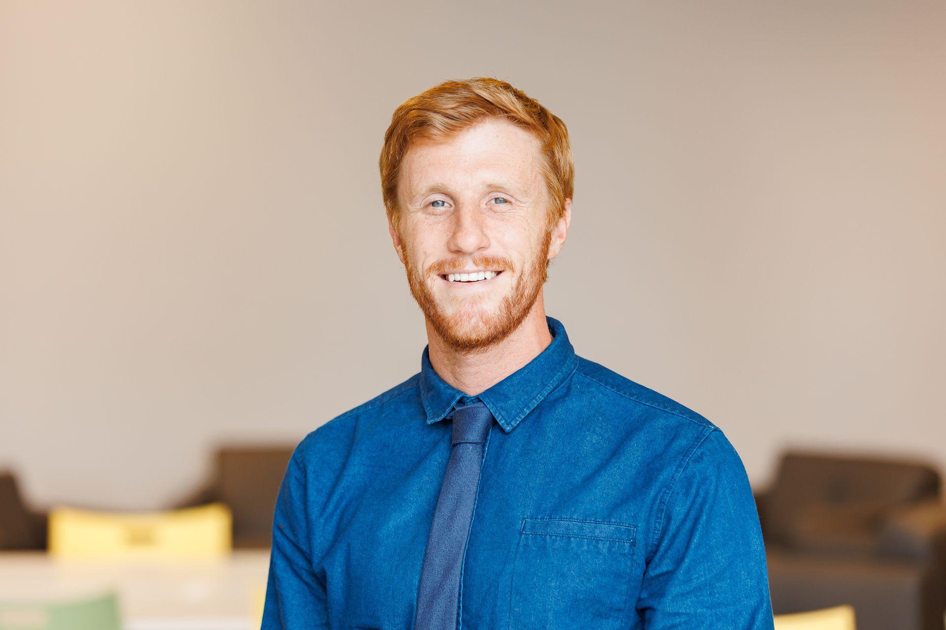 Smiling man with red hair and beard in a blue shirt and tie, standing indoors.